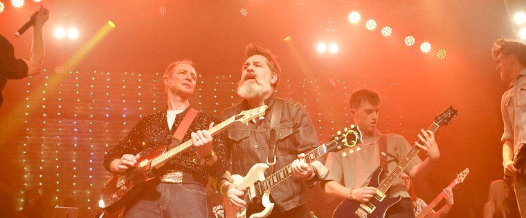Nick Robinson centre stage playing a white Gibson SG, flanked by other guitarists including a teenage player